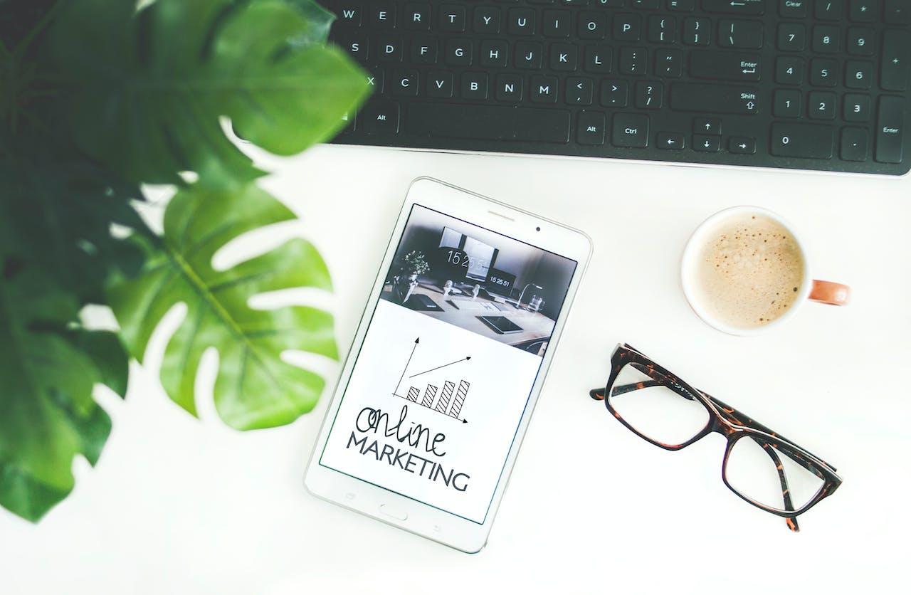 Online marketing displayed on a smartphone in front of laptop with a pair of glasses, coffee mug, and green plant